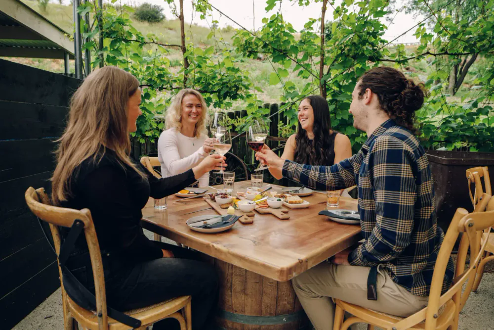 Four friends enjoying a meal and raising glasses of wine outdoors in a cozy restaurant garden surrounded by greenery.