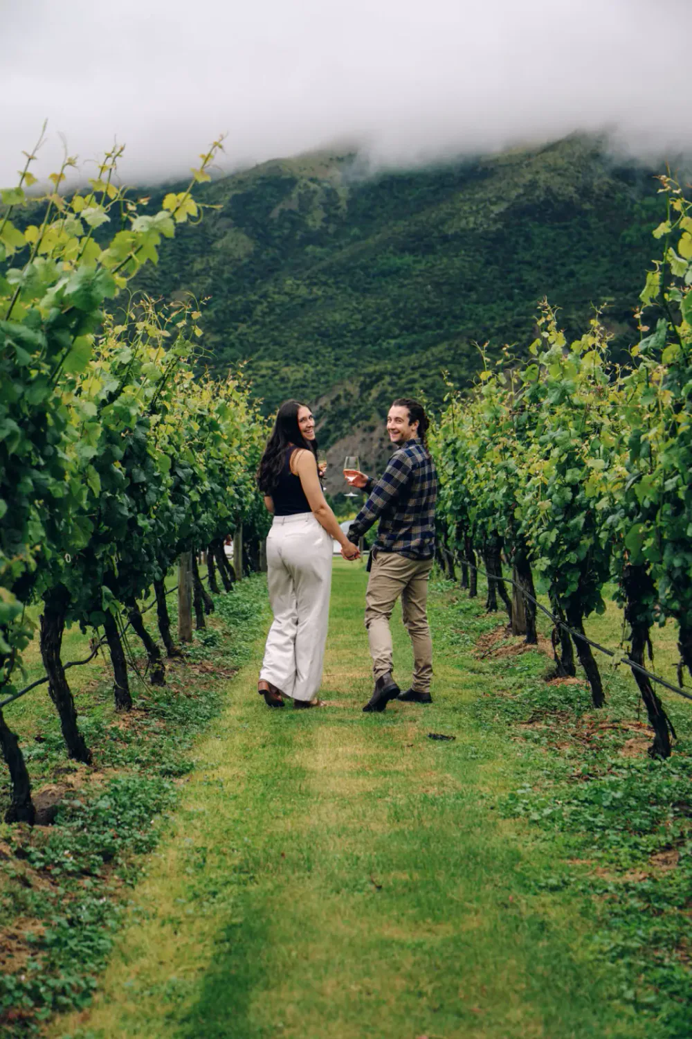 A couple holding hands and glasses of wine walking through a lush vineyard with green hills and misty mountains in the background.