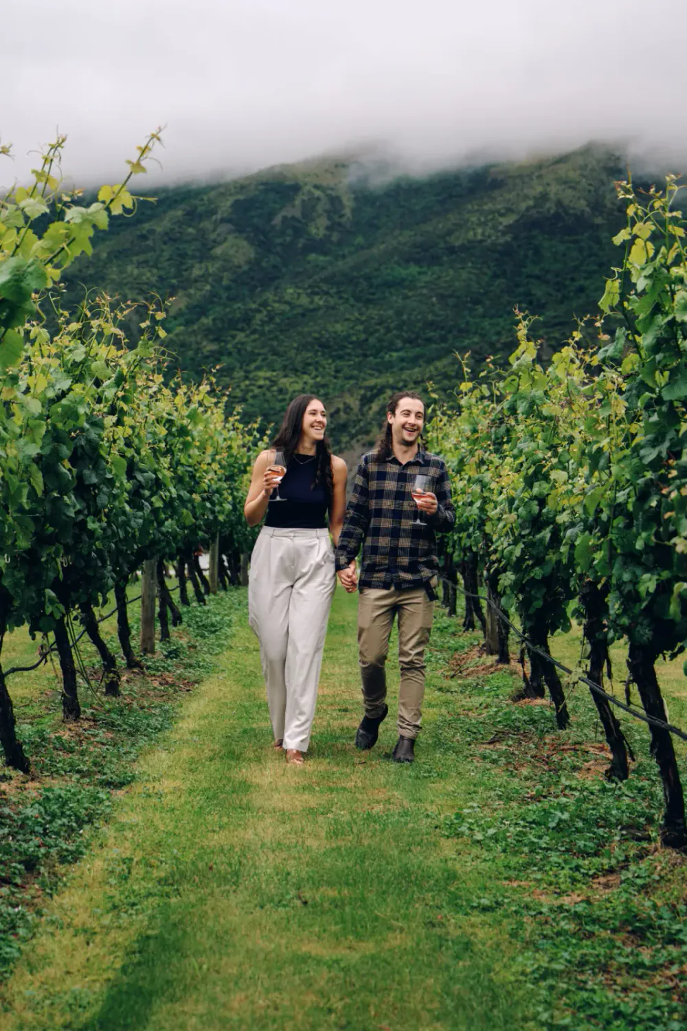 A couple walking hand in hand through a vineyard, holding glasses of wine and smiling, with green hills and cloudy sky in the background.