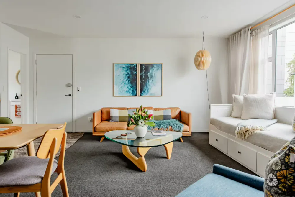 Living room with orange sofa, round glass coffee table with a flower arrangement, white daybed near window, artwork on wall, and a mix of modern and Scandinavian decor.