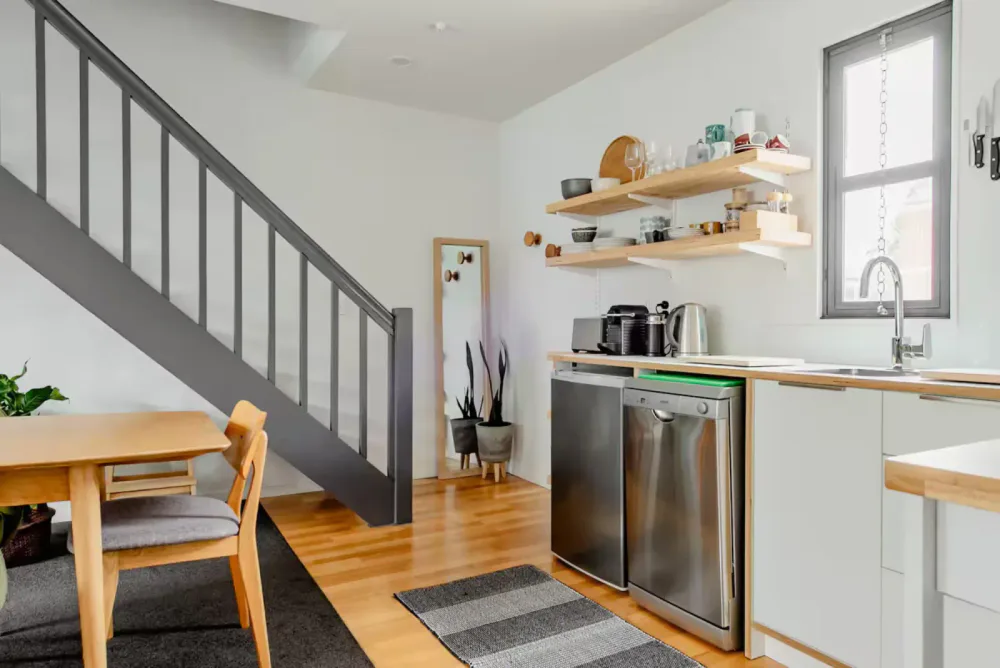 Kitchen area with white cabinets, wooden shelves holding dishes and glasses, a small window, a under-sink mini refrigerator, a coffee maker, kettle, and microwave on the counter, and a dining table with two chairs.