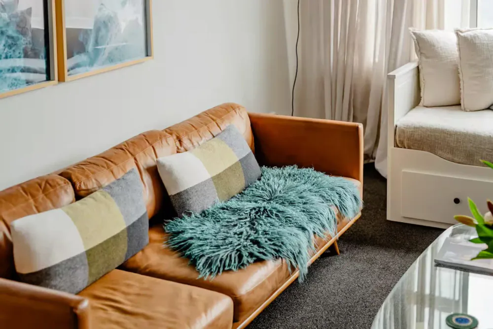 A living room with a tan leather sofa decorated with gray and green plaid pillows and a teal furry throw blanket, next to a white daybed with beige cushions, with sheer curtains in the background.