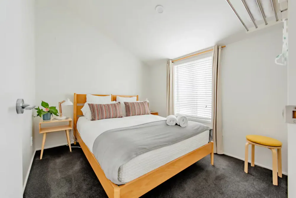 A guest bedroom prepared with fresh bedding, rolled towels, bedside lighting and a yellow stool.