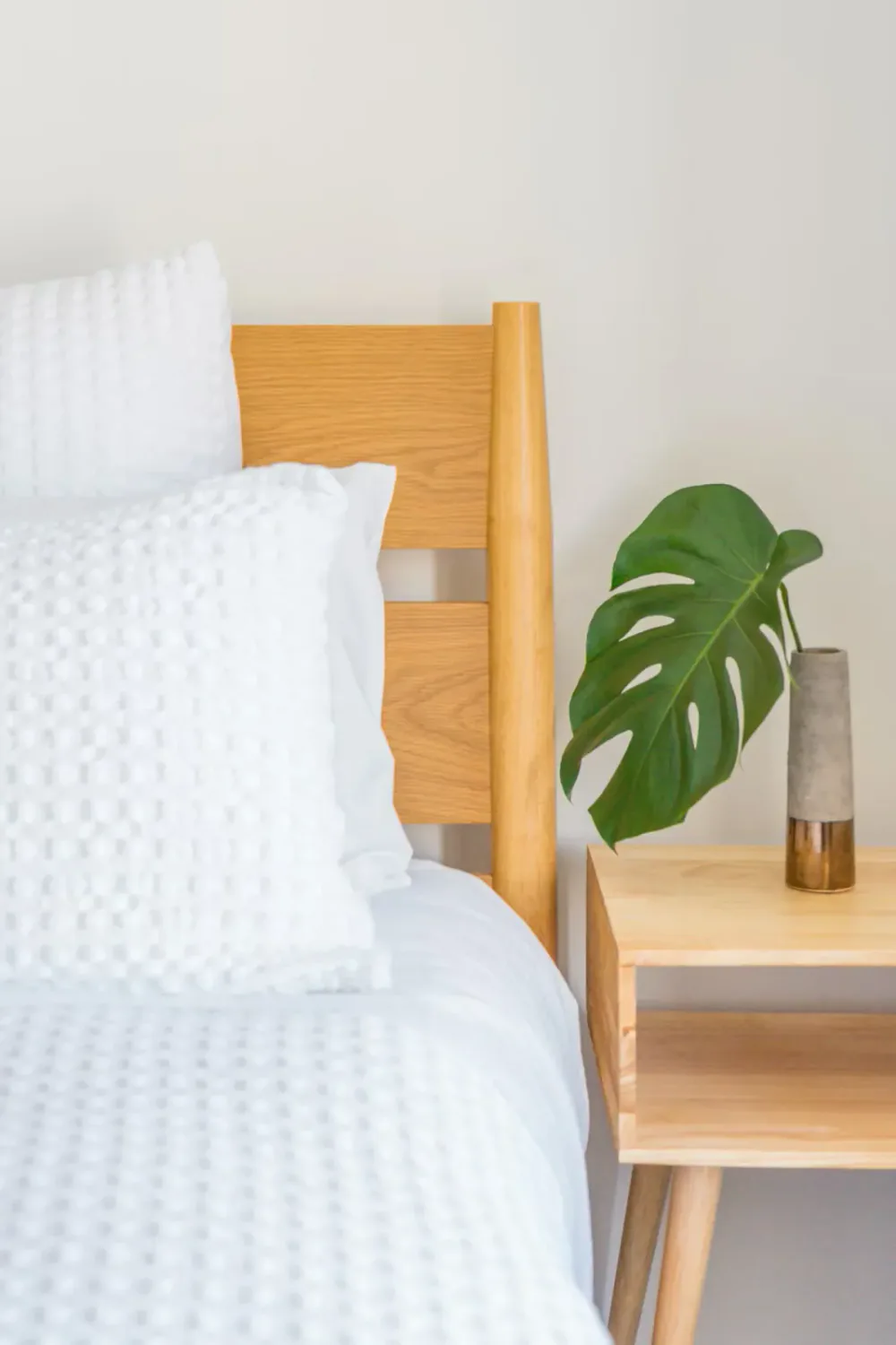 A bedroom with a wooden bed frame, white textured bedding and pillows, and a wooden nightstand with a green monstera leaf in a tall concrete and copper vase.