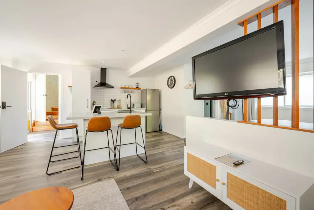 Modern open-concept kitchen and living room with wooden accents, a large flat-screen TV mounted above a white media console, bar stools at a kitchen island, stainless steel refrigerator, and a clock on the wall.