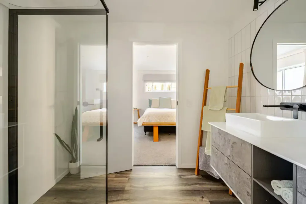 Modern bathroom with a view into a bedroom through a doorway, featuring a vanity with a sink, mirror, hanging towel rack, and a picture frame.