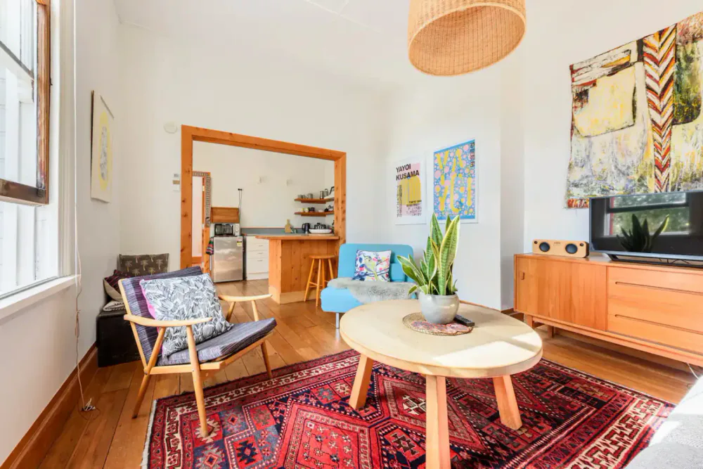 Living room with wooden furniture, colorful rugs, a potted plant on a coffee table, a blue armchair with a patterned pillow, and art on the walls. Open kitchen with countertop bar in background.