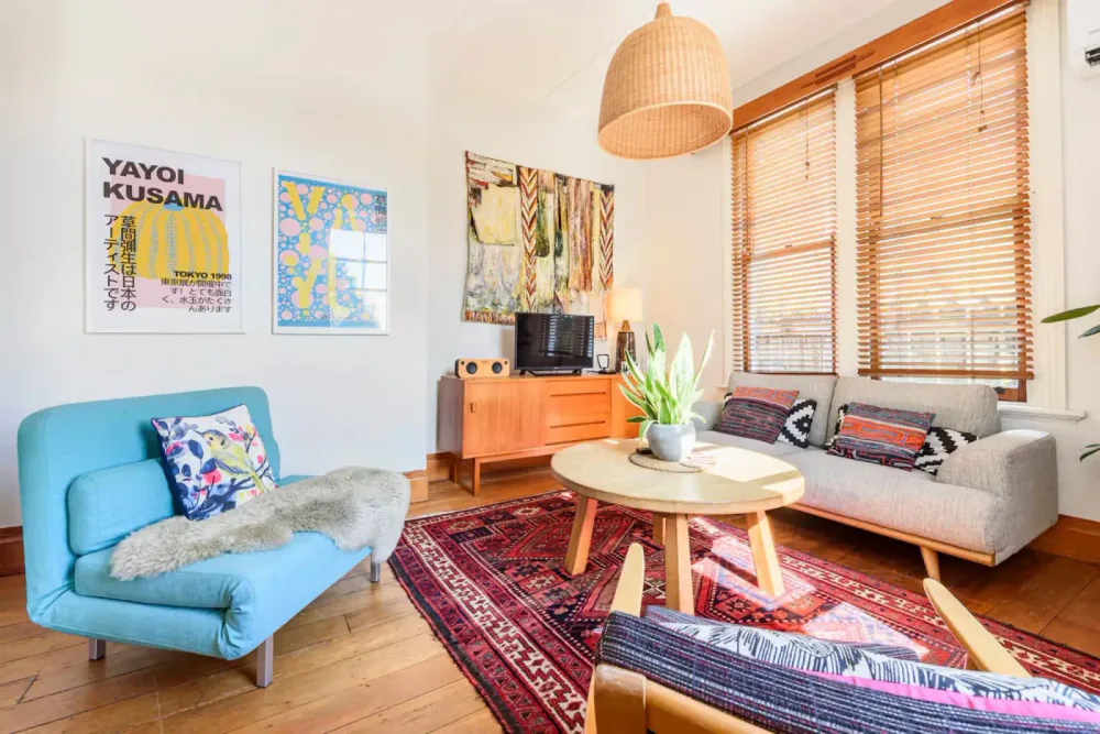 A bright living room with wooden blinds, relaxed seating, a red rug and mid-century furniture.