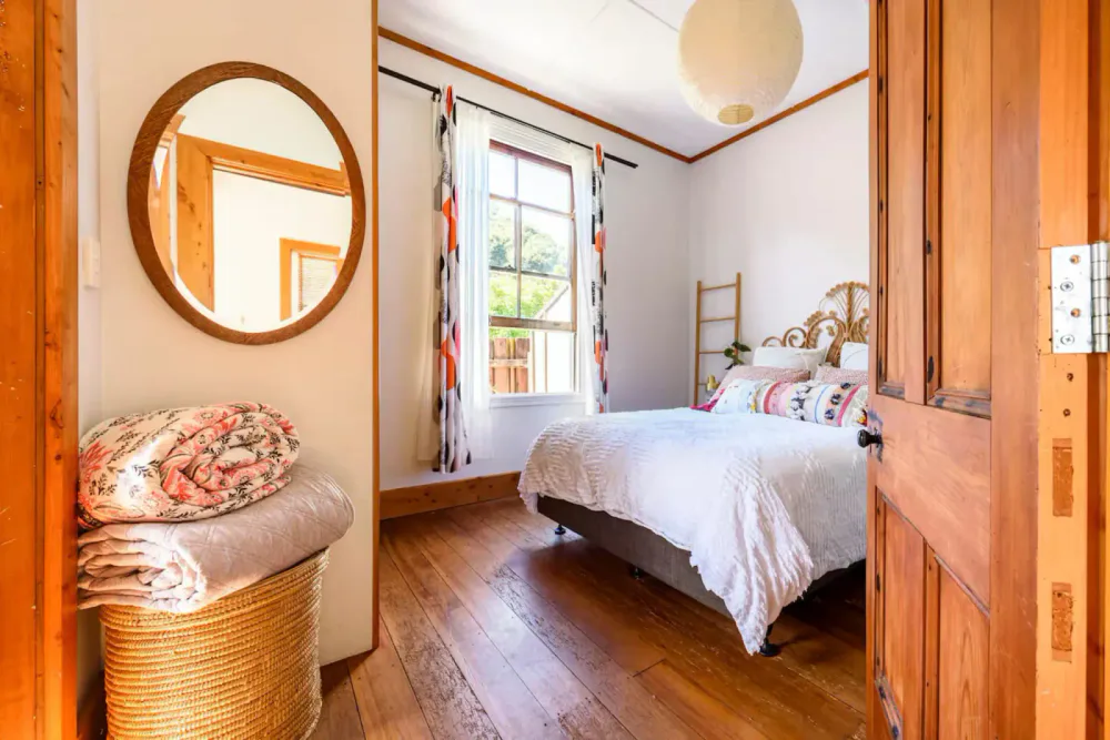 Cozy bedroom with wooden floors, a white bed with decorative pillows, a window with colorful curtains, a wicker basket with folded blankets, a round mirror on the wall, and a paper lantern ceiling light.