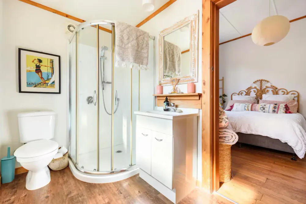 A bathroom with a corner shower, toilet, and a white sink vanity. A towel hangs above the shower. There is a framed artwork on the wall. The bathroom opens into a bedroom with a bed and wooden flooring.