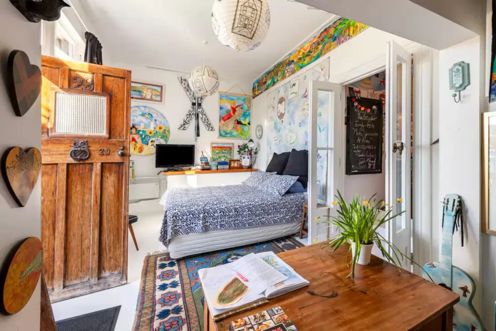 A cozy bedroom with colorful artwork on the walls, a wooden door, a bed with patterned bedding, a small desk with a TV, potted plant on a wooden table, and various decorations and furniture.