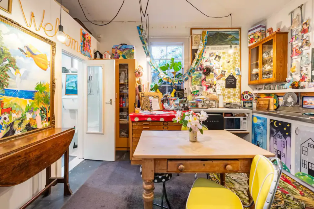 Colorful, cluttered kitchen and dining area with various decorations, paintings, and plants, featuring a wooden table with a vase of flowers, yellow chairs, a red tablecloth, and a window with sunlight.
