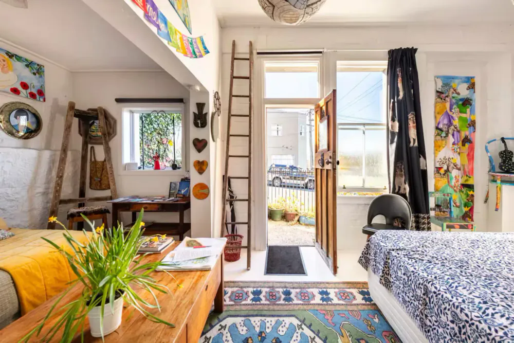 Interior of a cozy room with a bed on the right, a wooden table with a potted plant on the left, an open front door leading outside, and various colorful decorations on the walls.