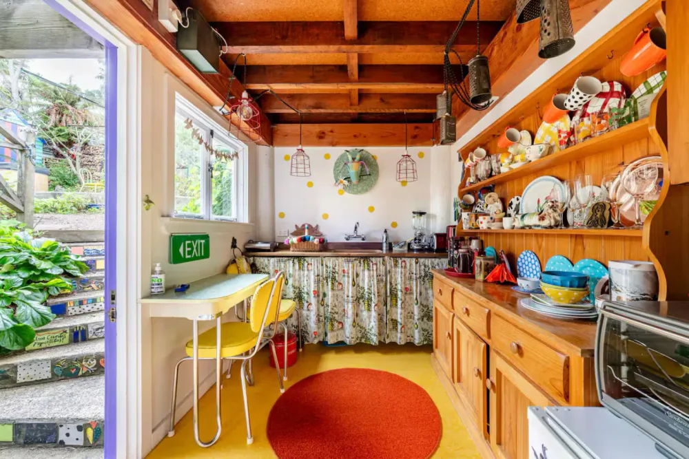 Colorful kitchen with wooden shelves and ceiling beams, yellow chairs, a green exit sign, a window with outside greenery, and various dishes and kitchen appliances.