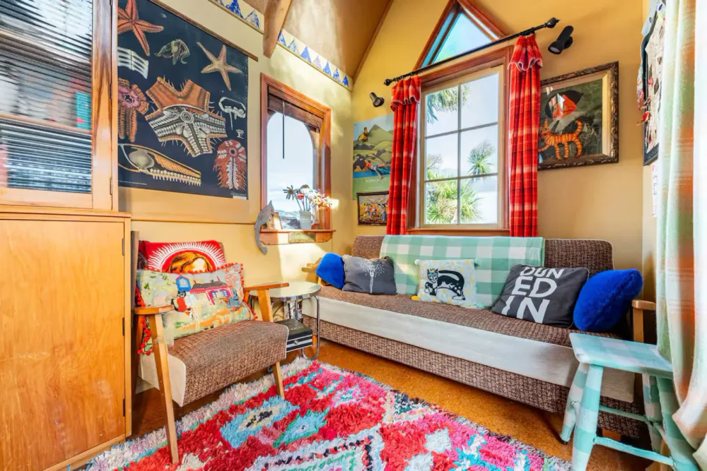 Cozy living room with colorful decor, wooden furniture, and sea life art on the walls. There are two windows with red curtains and a patterned rug on the floor.
