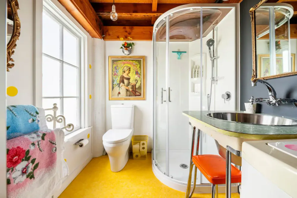 Bathroom with a toilet, shower enclosure, sink, mirror, window, framed artwork, and colorful decor including towels and a yellow floor.