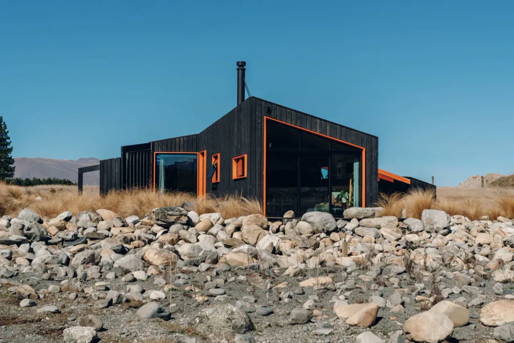 Modern black house with orange accents sitting on rocky terrain, surrounded by dry grass under a clear blue sky.