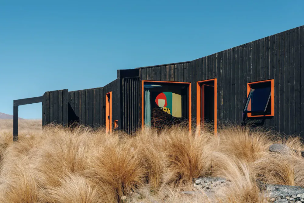 Contemporary black wooden house with orange window frames surrounded by dry grass under a clear blue sky.