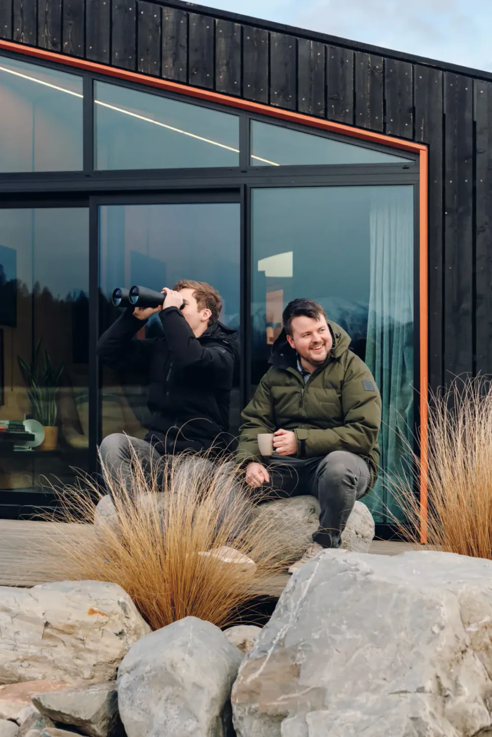 Two men sitting outside a modern house on rocks surrounded by dry grasses, one looking through binoculars, the other holding a mug, smiling.