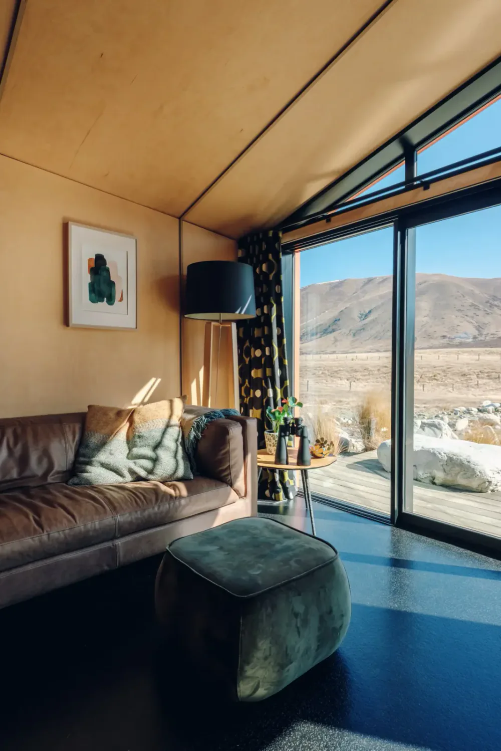 Modern living room with wooden walls, a leather sofa with pillows, a small side table with binoculars and potted plants, a large black floor lamp, and floor-to-ceiling windows showing a mountainous landscape and blue sky outside.