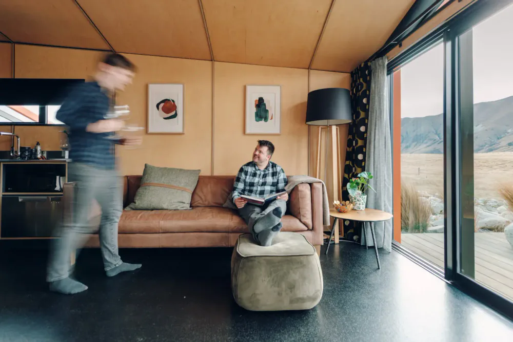 A guest reads on a leather sofa in a warm timber living room while another person walks past.