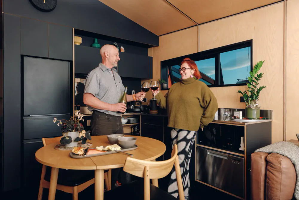 A man and woman clinking glasses of red wine in a modern kitchen, with a wooden dining table and a plate of cheese and crackers in the foreground.