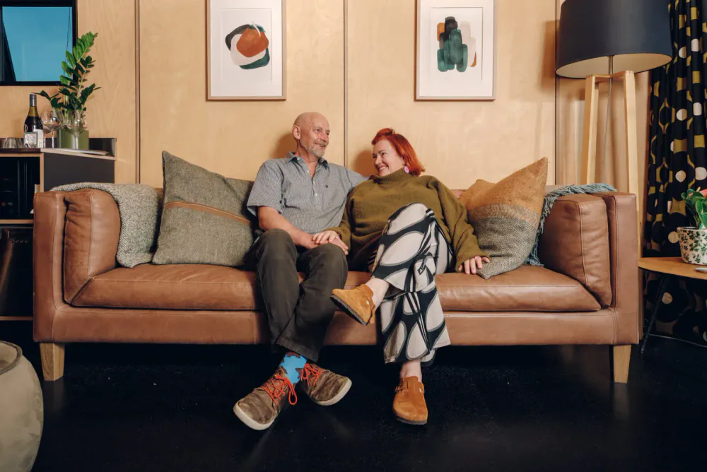 A man and a woman sitting on a brown leather couch, holding hands and smiling at each other, in a cozy living room with abstract artwork, a table lamp, and decorative pillows.