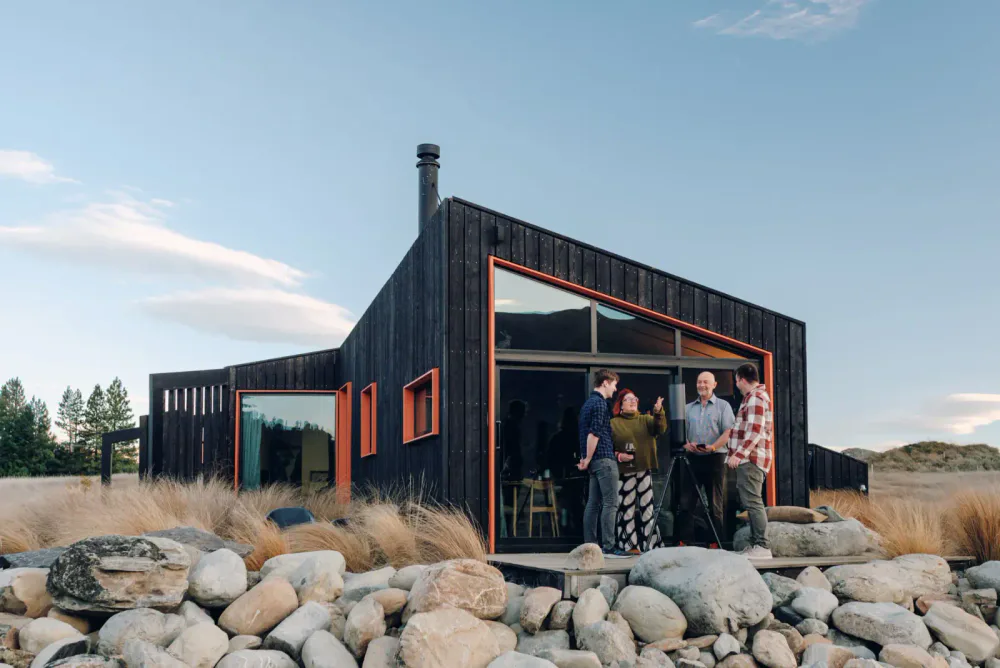 Group of four people standing on a deck outside a modern black house with large glass windows, talking and smiling, with rocks and tall grasses in the foreground and trees in the background.