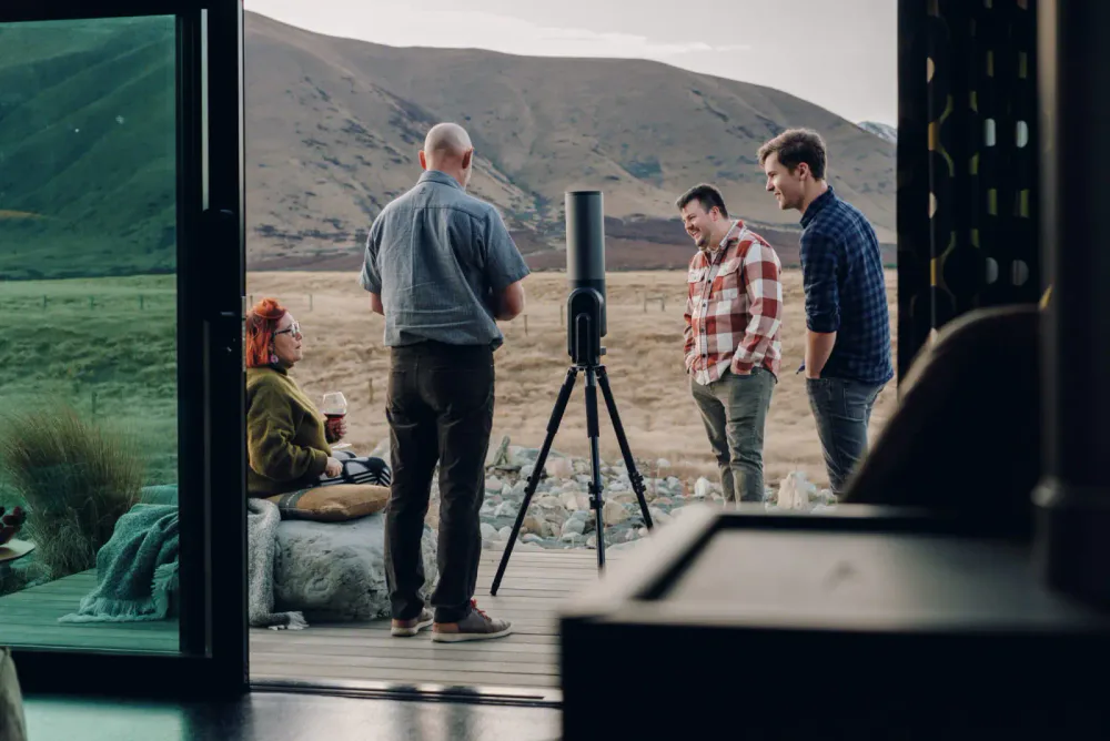 Four people stand outside on a deck in a rural, mountainous landscape, with one woman sitting on a rock holding a glass of red wine, while the three men talk nearby, with a large black speaker on a tripod set between them.