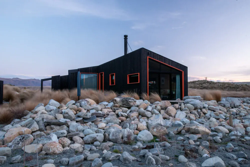 Modern black house with orange trim in a rocky, desert landscape under a partly cloudy sky.