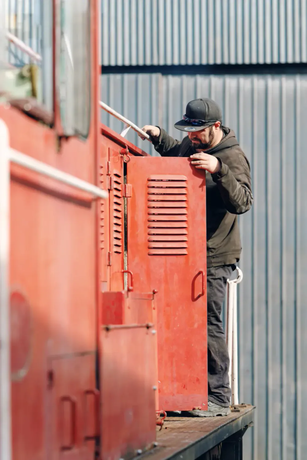 A man in a black hoodie, black cap, and sunglasses is working on an orange industrial machine or cabinet outdoors with a metal wall in the background.