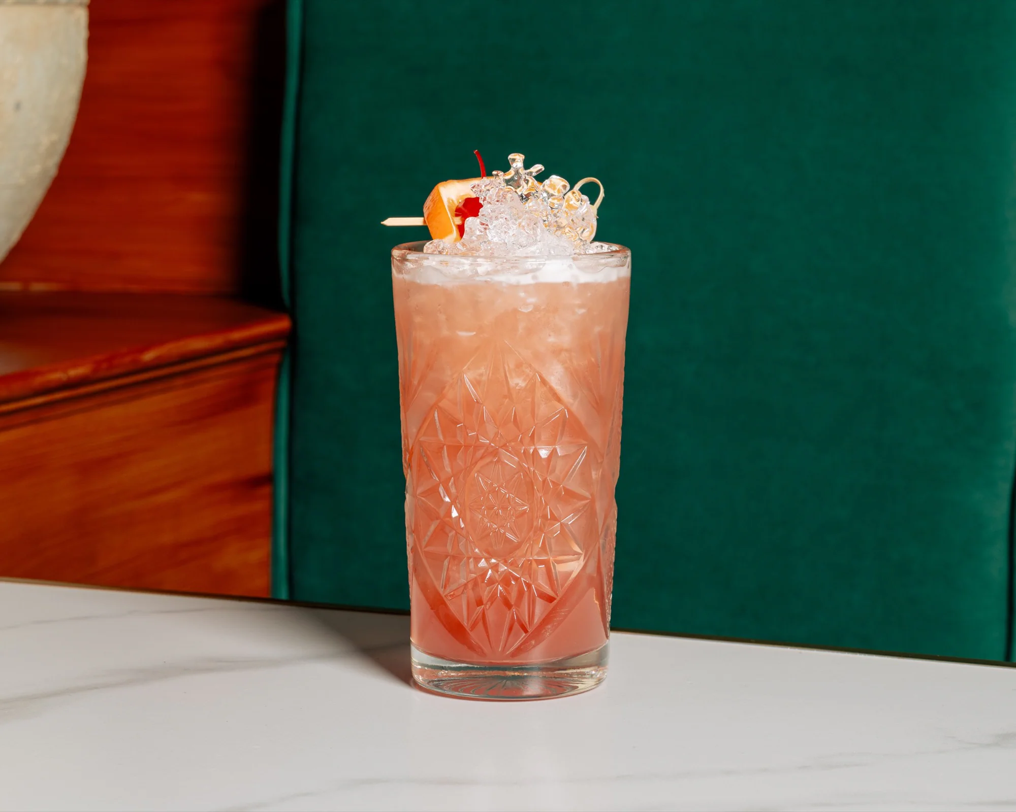 A pink cocktail with citrus garnish and crushed ice against a green booth