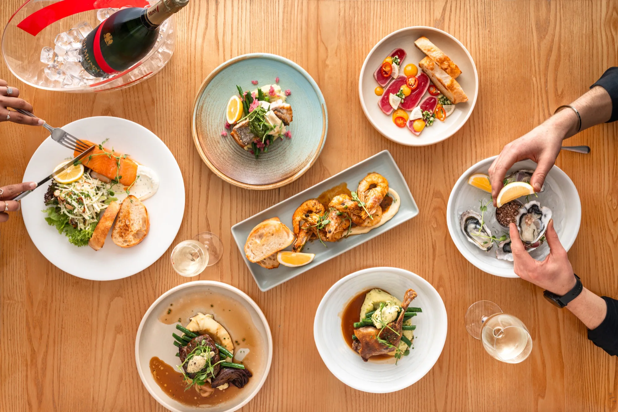 An overhead restaurant table set with seafood, meat dishes, wine and diners’ hands
