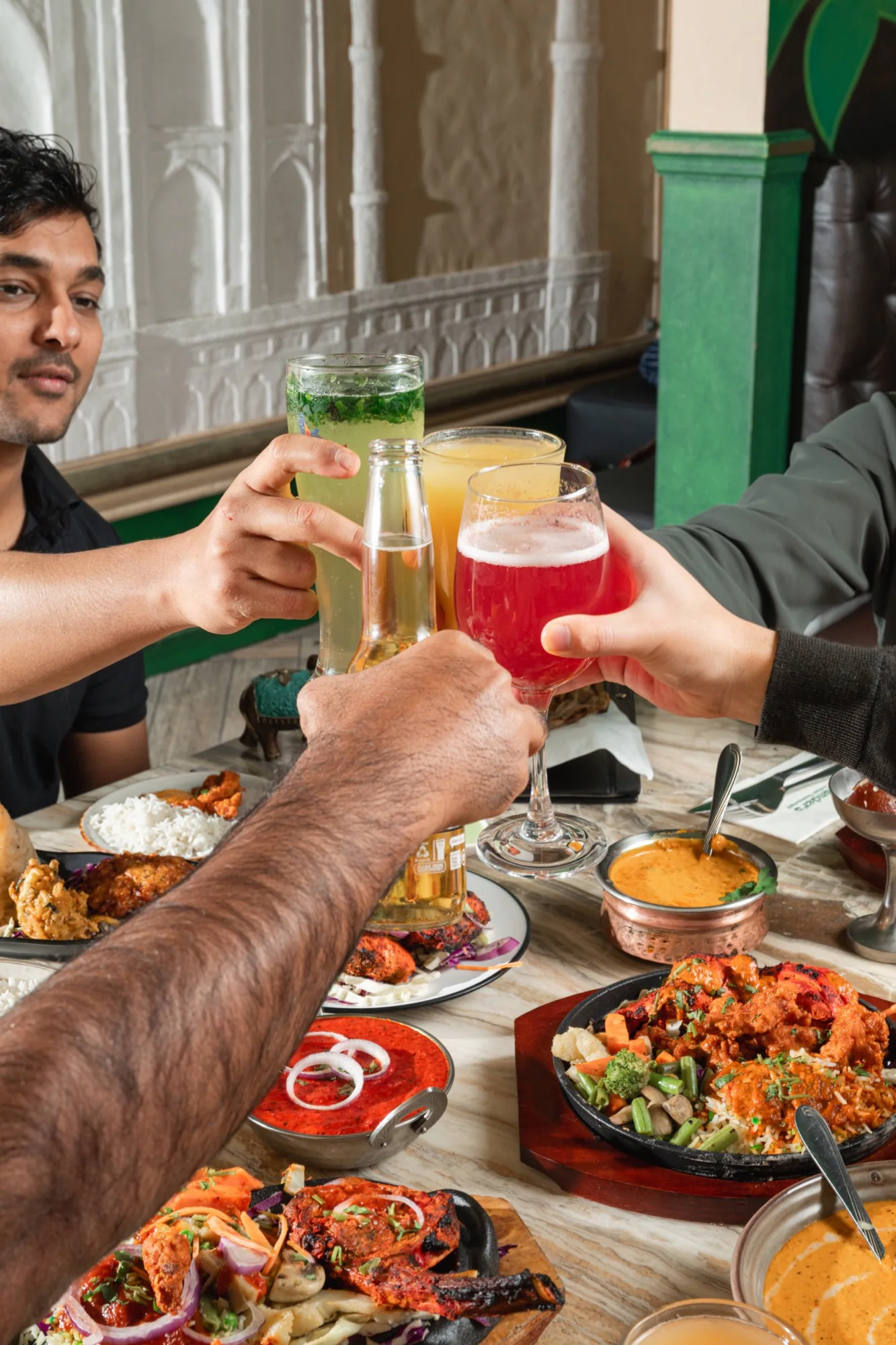Restaurant guests raising colourful drinks above a table of shared Indian dishes