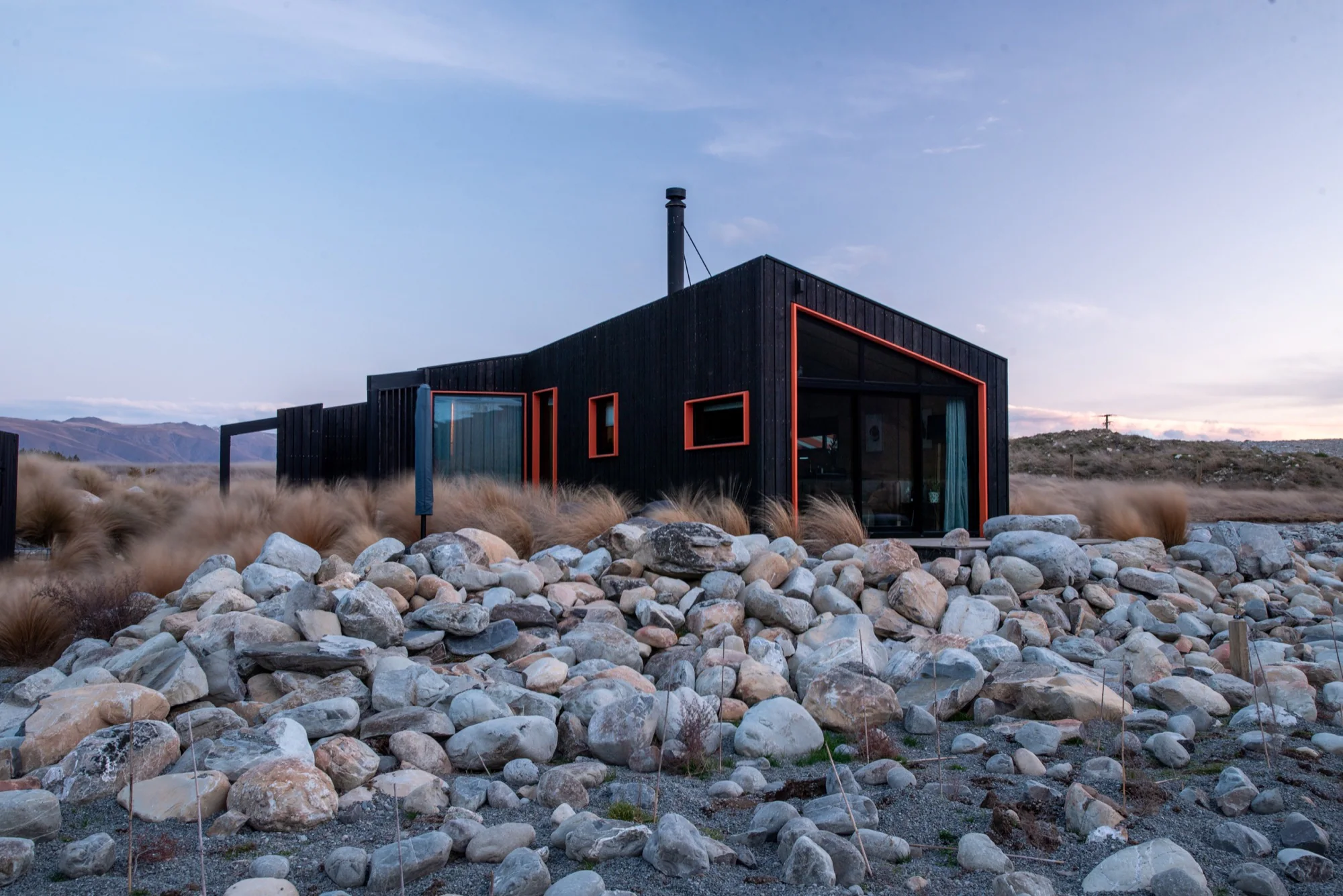 A black timber cabin with orange window frames among rocks and tussock at dusk