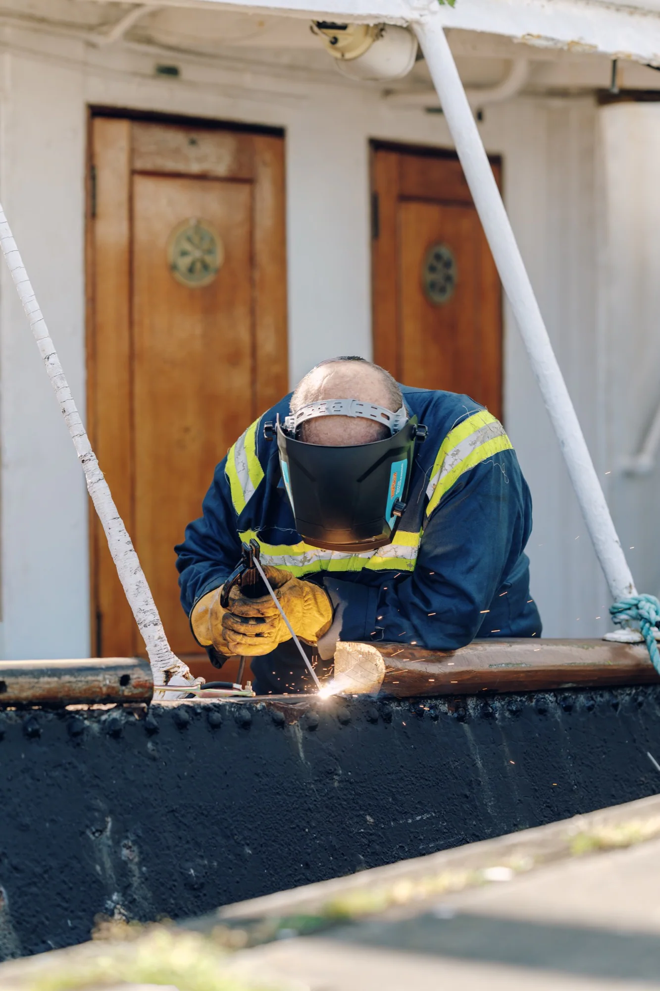 A worker wearing a welding helmet, gloves and high-visibility workwear welding a metal rail outdoors